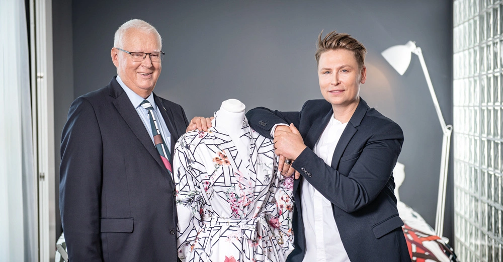 Zwei Männer in Anzügen stehen neben einer Schneiderpuppe mit einem gemusterten Kleidungsstück in einem modern eingerichteten Raum mit grauen Wänden und einer weißen Stehlampe im Hintergrund