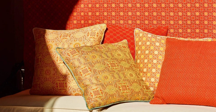 Kissen liegen auf einem cremefarbenen Sofa vor einer Wand mit rot-gelbem geometrischem Muster und werden von Sonnenlicht beleuchtet.