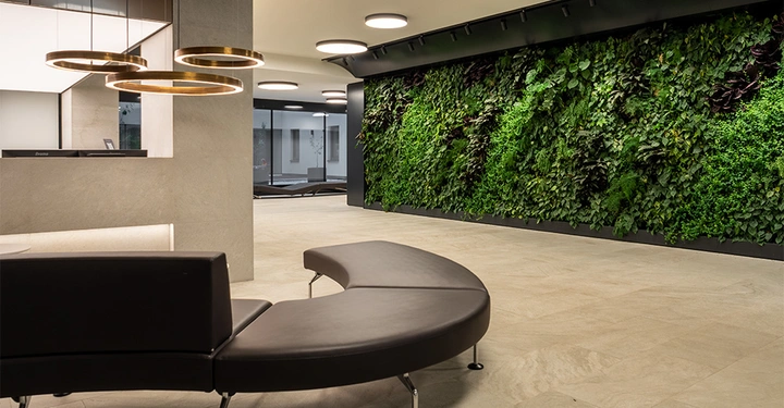 Galerie-Bild 3: Halbrundes, schwarzes Sofa steht vor einer Wand mit üppigem grünem Pflanzenbewuchs in einem modernen, gut beleuchteten Empfangsbereich. Im Hintergrund befindet sich ein Empfangstresen mit hängenden Leuchten.