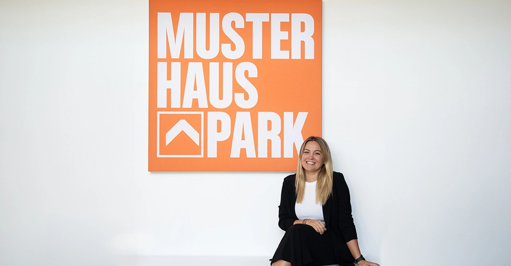 Frau sitzt lächelnd vor einem großen orangefarbenen Schild in einem minimalistischen Raum. Auf dem Schild steht Muster Haus Park mit einem stilisierten Pfeilsymbol darunter.