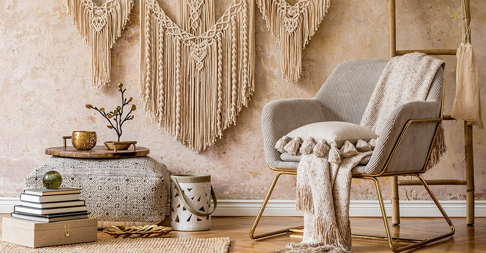 Gemütliche Wohnzimmerecke im Kolonialstil mit Makramee-Wanddeko, Sessel mit Kissen und Decke, orientalischem Pouf, Dekotablett und Naturfarben. 