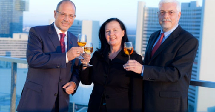 Galerie-Bild 12: Drei Personen in formeller Kleidung halten Sektgläser und stoßen an auf einer Dachterrasse mit modernen Hochhäusern im Hintergrund bei Tageslicht.