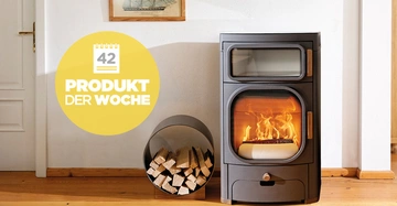 Schwarzer Kaminofen brennt im Wohnzimmer neben einem Metalkorb mit Holzscheiten an einer weißen Wand mit einem Bild. Text: Produkt der Woche 42
