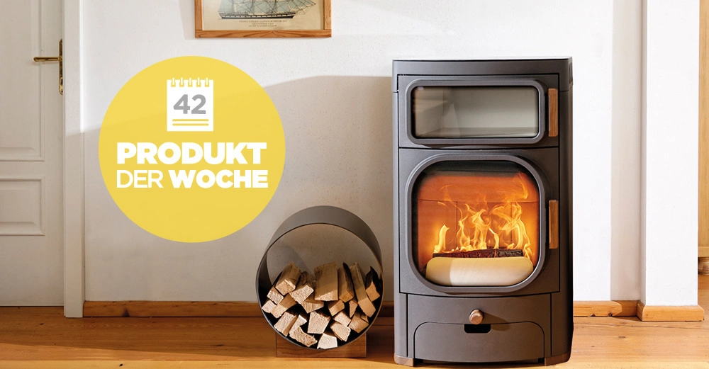 Schwarzer Kaminofen brennt im Wohnzimmer neben einem Metalkorb mit Holzscheiten an einer weißen Wand mit einem Bild. Text: Produkt der Woche 42