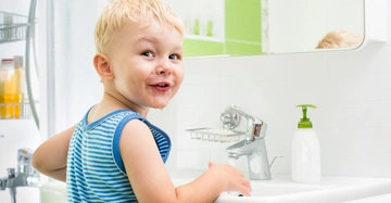 Kleinkind lacht während es sich die Hände an einem Waschbecken in einem hell erleuchteten Badezimmer wäscht.aign