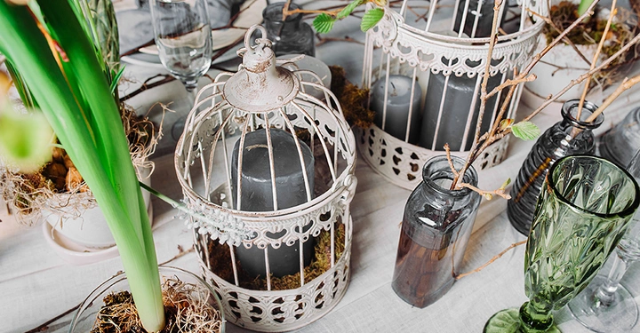 Mehrere dekorative Vogelkäfige enthalten Kerzen und Moos auf einem Tisch mit Glasflaschen und grünen Weingläsern im Vordergrund umgeben von Pflanzen und Ästen.