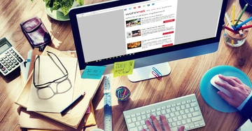 Person tippt am Computer in einem kreativen Büro mit Pflanzen, Brillen, Notizen, Stiften, Taschenrechner und Schreibtischzubehör. Text auf dem Monitor: wohnnet WOHNEN auf einem Blog. Notizen enthalten "IDEE: ACTION PLAN" und "TEI 101: 3 Email Check".