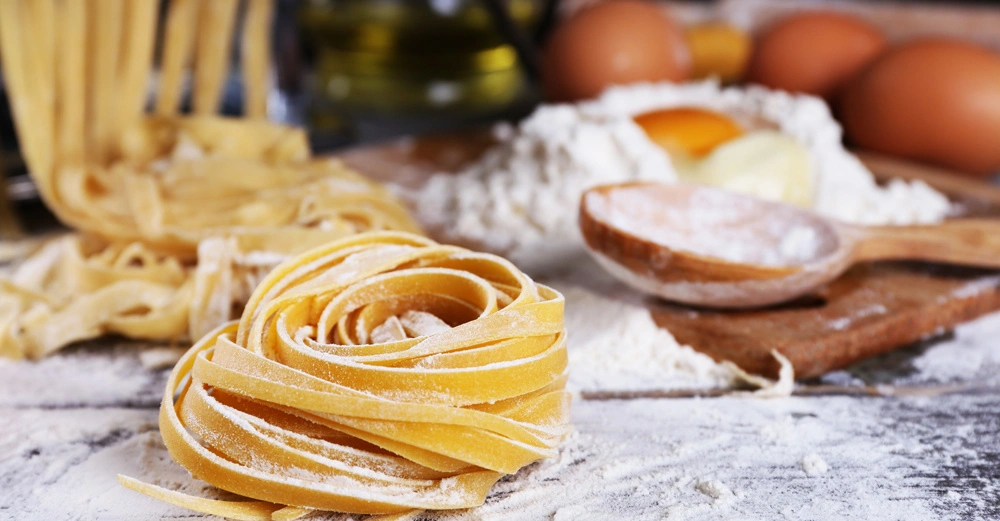Hausgemachte Nudeln, die auf bemehlter Oberfläche liegen; im Hintergrund befinden sich Eier, Mehl und ein Holzlöffel; eine Küchenszene.