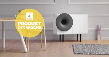 Weiße Kommode mit großem grauem Lautsprecher; steht im Wohnzimmer neben einem Tisch und einem Teppich; Text im gelben Kreis: 4 Produkt der Woche.