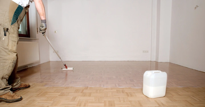Eine Person lackiert den Holzboden mit einer Rolle in einem leeren Raum; daneben steht ein weißer Kanister.