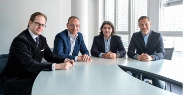 Vier Männer in Anzügen sitzen an einem geschwungenen Besprechungstisch in einem modernen Büro mit weißen Wänden und großen Fenstern im Hintergrund