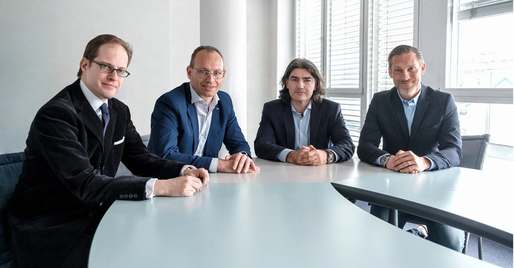 Vier Männer in Anzügen sitzen an einem geschwungenen Besprechungstisch in einem modernen Büro mit weißen Wänden und großen Fenstern im Hintergrund