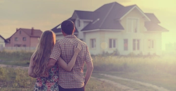 Ein Paar steht Arm in Arm in einem Feld und blickt auf ein weißes Einfamilienhaus im Hintergrund bei Sonnenuntergang.