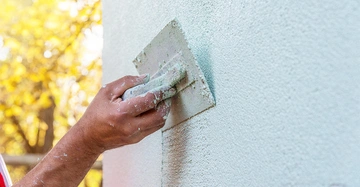männliche Hand mit Spatel bringt weißen Rauhputz an Fassade an, Großaufnahme mit herbstlichem Baum und Sonnenschein im Hintergrund.