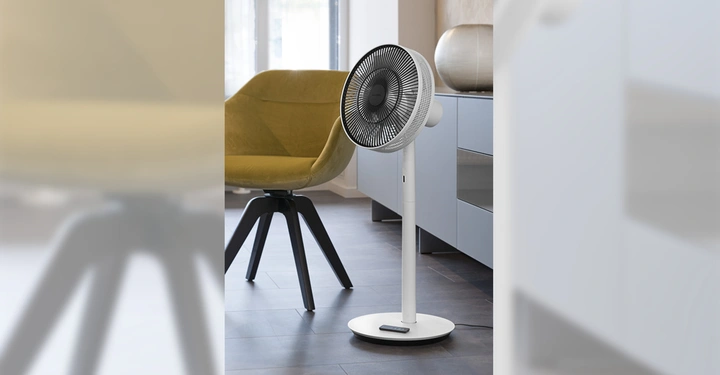 Ventilator steht auf dem Boden in einem modernen Wohnzimmer vor einem gelben Sessel und einer grauen Kommode mit dekorativer Vase und Fenster im Hintergrund.