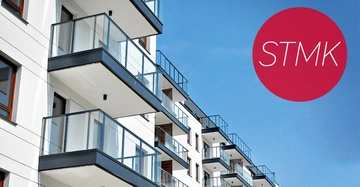 Mehrere moderne Wohngebäude mit Balkonen stehen nebeneinander unter einem klaren blauen Himmel. Ein roter Kreis mit weißem Text "STMK" ist am rechten Rand zu sehen.