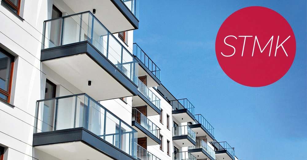 Mehrere moderne Wohngebäude mit Balkonen stehen nebeneinander unter einem klaren blauen Himmel. Ein roter Kreis mit weißem Text "STMK" ist am rechten Rand zu sehen.