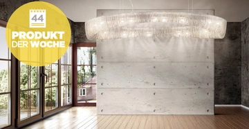 Ein großer Kronleuchter hängt von der Decke vor einer Betonwand in einem Raum mit Holzboden und großen Fenstern. Text im gelben Kreis: 44 PRODUKT DER WOCHE.