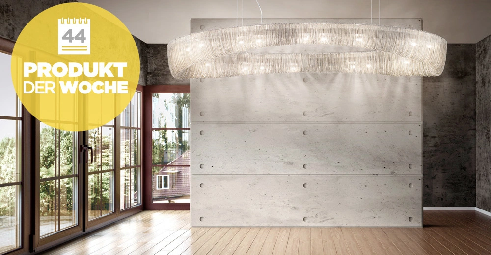 Ein großer Kronleuchter hängt von der Decke vor einer Betonwand in einem Raum mit Holzboden und großen Fenstern. Text im gelben Kreis: 44 PRODUKT DER WOCHE.