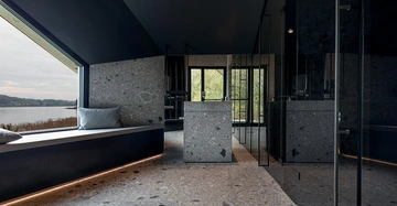 Sitzbank mit Kissen neben einem großen Fenster mit Seeblick moderne graue Steinelemente und Spiegel im Raum große Fenster in einem eleganten Innenraum mit heller und grüner Außenansicht