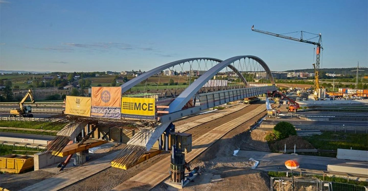 Galerie-Bild 6: Eine im Bau befindliche Brücke wird von Kränen und Arbeitern auf einer Baustelle über Autobahnen montiert. Drei große Schilder tragen die Aufschriften MCE Haslinger und Rhomberg.