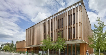 Modernes dreistöckiges Gebäude mit Holzfassade und vertikalen Lamellen Fenster. Stehende Bäume und Fahrräder im Vordergrund. Im städtischen Umfeld. Text: Verein für Baukultur - Türkenschanz.