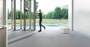 Person öffnet eine große Falttür in einem modernen, weißen Raum mit Blick auf einen grünen Garten mit Bäumen. Ein kleiner weißer Hocker steht rechts im Raum.