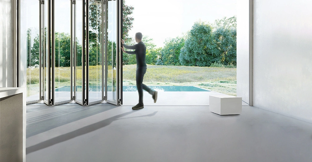 Person öffnet eine große Falttür in einem modernen, weißen Raum mit Blick auf einen grünen Garten mit Bäumen. Ein kleiner weißer Hocker steht rechts im Raum.