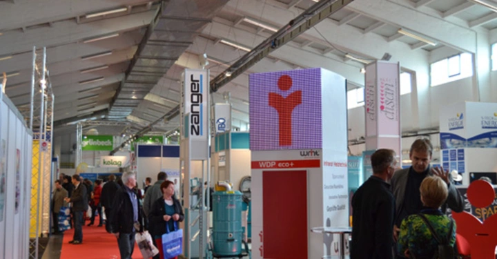 Galerie-Bild 7: Menschen schlendern durch eine Messehalle mit Ständen von verschiedenen Unternehmen. In der Mitte ein roter Stand mit einem stilisierten Menschenlogo. Beschilderungen der Firmen Zanger und Kaibach sichtbar.