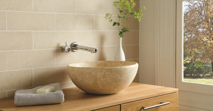 Galerie-Bild 7: Waschbecken aus Stein steht auf einer Holzplatte eingebaut in ein Badezimmer mit beigen Fliesen ausgelegt auf der Rückwand und Draufsicht auf herbstliche Landschaft durch ein großes Fenster.