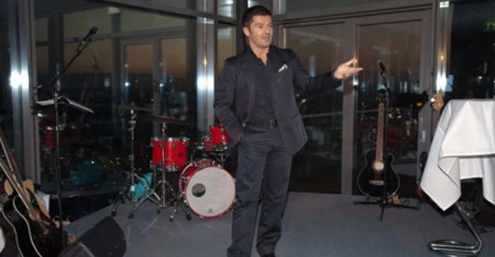 Galerie-Bild 65: Mann spricht vor Publikum auf Bühne in einem Raum mit Glaswänden bei Nacht Hinter ihm Musikinstrumente wie Gitarren und ein Schlagzeug aufgestellt.