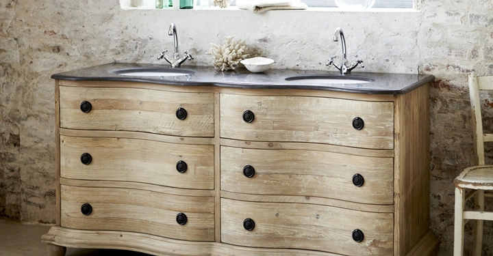 Galerie-Bild 5: Holzkommode mit geschwungenen Schubladen stehen in einem rustikalen Badezimmer. Zwei Waschbecken mit Wasserhähnen befinden sich darauf. Ein Fenster im Hintergrund beleuchtet den Raum.