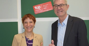 links kleinere Frau mit kurzen roten Haaren, rechts großer Mann mit Brille und grauem Haar, beide im Businesslook, lächeln in die Kamera.
