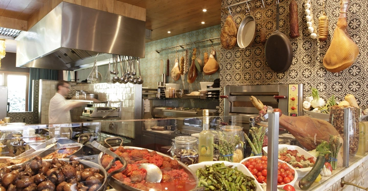 Galerie-Bild 5: Koch bereitet hinter Glasvitrine Speisen zu Küche im traditionellen Stil dekoriert mit hängenden Würsten Pfannen Kräutern Schalen mit Fleisch Gemüse Weinen Herd und Ofen an der Wand.