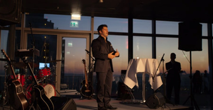 Galerie-Bild 69: Ein Mann steht auf einer Bühne und spricht in ein Mikrofon; die Bühne ist mit Instrumenten ausgestattet. Im Hintergrund sind große Fenster, durch die man einen Sonnenuntergang und eine Stadtlandschaft sieht.