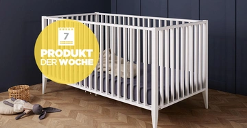 Ein weißes Babybett steht auf einem Holzboden in einem Raum mit dunklen Wänden. Links liegt ein Stoffspielzeug neben einem Korb. Text im gelben Kreis: "7 PRODUKT DER WOCHE".