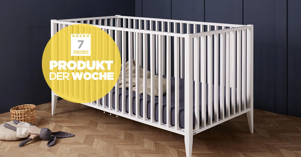 Ein weißes Babybett steht auf einem Holzboden in einem Raum mit dunklen Wänden. Links liegt ein Stoffspielzeug neben einem Korb. Text im gelben Kreis: "7 PRODUKT DER WOCHE".