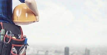 Bauarbeiter hält gelben Schutzhelm trägt Werkzeuggürtel mit verschiedenen Werkzeugen Hintergrund zeigt verschwommene Stadtlandschaft