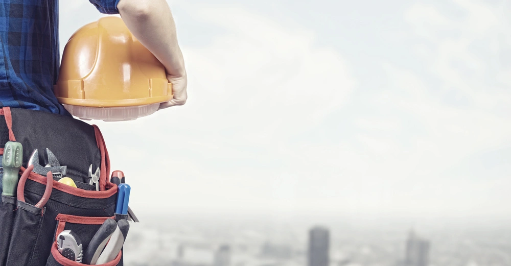 Bauarbeiter hält gelben Schutzhelm trägt Werkzeuggürtel mit verschiedenen Werkzeugen Hintergrund zeigt verschwommene Stadtlandschaft