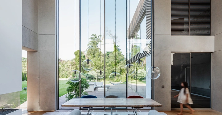 Galerie-Bild 1: Großer Holztisch steht vor raumhohen Fenstern welche auf einen grünen Garten mit Terrasse blicken. Modernes Interieur mit Hängelampen. Kind läuft rechts durch die Wohnung.