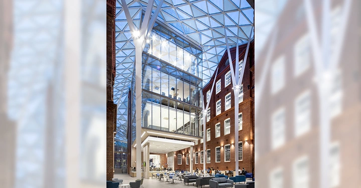 Galerie-Bild 3: Ein moderner Glasgebäudekomplex steht in einem Innenhof mit hohen roten Backsteinwänden und einem Gitterdach. Menschen sitzen und unterhalten sich in einer hell erleuchteten Sitzbereich.