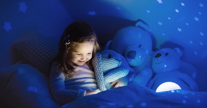 Galerie-Bild 2: Mädchen lächelt und hält Stofftier im Bett umgeben von zwei Kuschelbären in einem blau beleuchteten Raum mit leuchtenden Sternprojektionen.