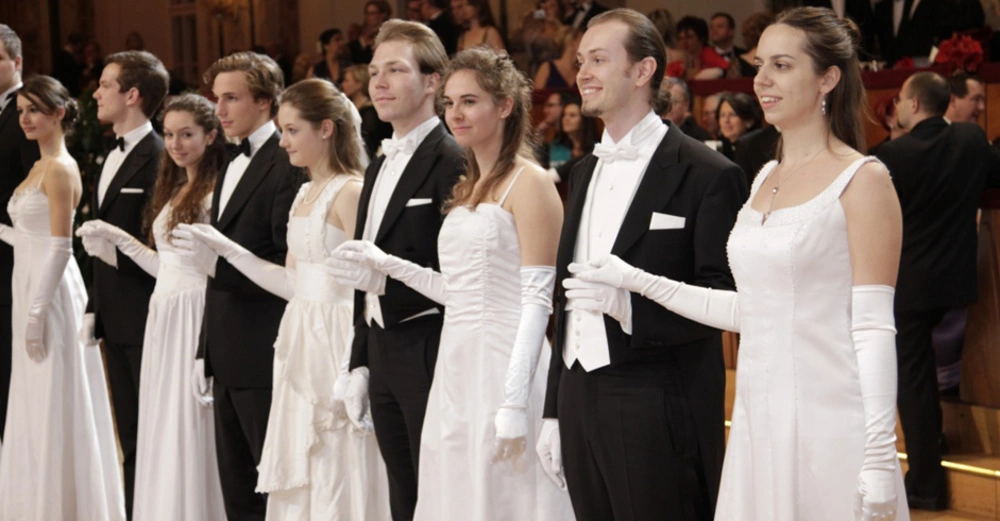Eine Gruppe junger Menschen in festlicher Kleidung steht in einer Reihe Händchen haltend in einem eleganten Saal mit Menschen im Hintergrund.