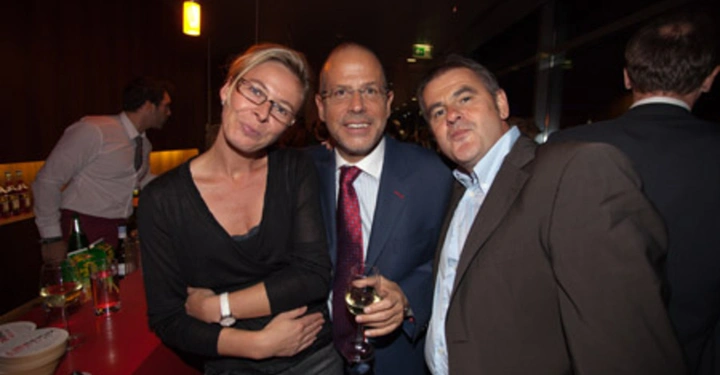 Galerie-Bild 83: Eine Frau und zwei Männer posieren lächelnd für ein Foto in einem gedämpft beleuchteten Raum während einer gesellschaftlichen Veranstaltung mit Getränken auf einer roten Theke im Hintergrund.