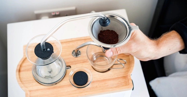 Hand hält ein Sieb mit Kaffeepulver über eine Kaffeemaschine auf einem Holztablett auf einem weißen Tisch in einer Küche.