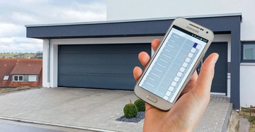 Hand hält Smartphone Display zeigt Steuerungs-App Einfahrt moderner Garage im Hintergrund Haus mit Flachdach und Pflastersteinen Umgebung ruhige Wohngegend mit roten Dächern und Natur im Hintergrund