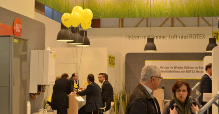 Galerie-Bild 17: Männer sitzen an einem Standtisch und unterhalten sich mit Beratern in einem modernen Messestand mit Heizungs- und Klimaanlagen. Ein Schild trägt die Aufschrift Heizen mit Sonne Luft und ROTEX.