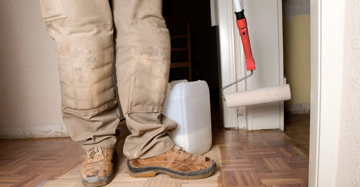 Beine einer Person in Arbeitshosen und Arbeitsstiefeln stehen auf einem Holzboden während sie eine Farbrolle und einen weißen Plastikkanister halten in einem Raum mit gelbem und weißem Anstrich.