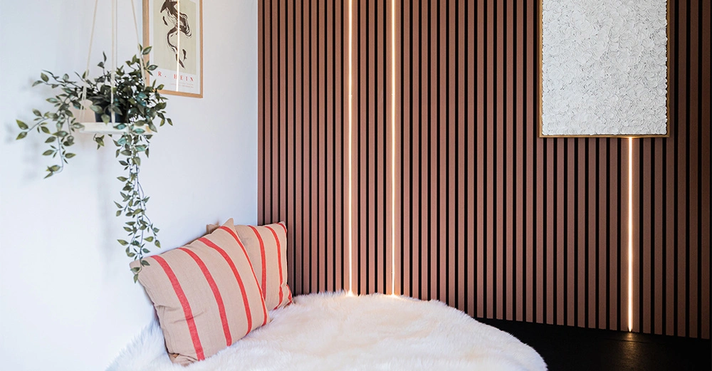 Ein weiches weißes Fell liegt in einer Ecke vor einer holzvertäfelten Wand zwei rot gestreifte Kissen Pflanzen im Hängekorb und zwei gerahmte Bilder an der Wand.