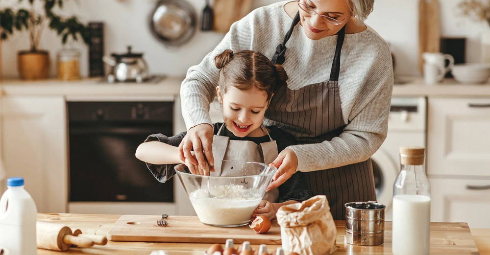 Oma und Enkelin stehen in der Küche und bereiten einen Kuchenteig zu. Holzarbeitsplatte, im Hintergrund helle Küchenmöbel und Pfannen an der Wand. Fröhliche Stimmung.